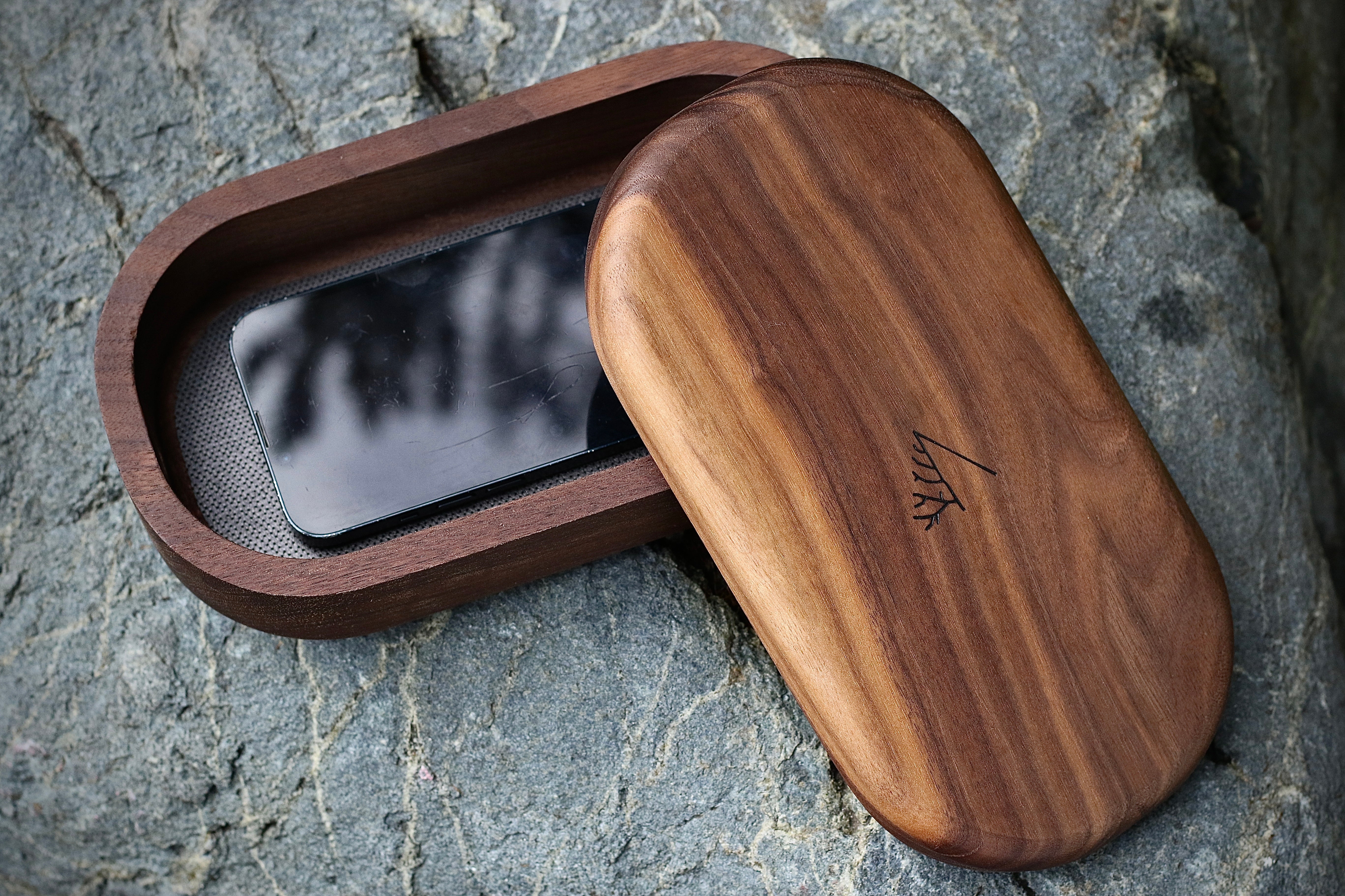 Phone Tomb in walnut, open on grey stone, a phone lying inside and the lid beside it